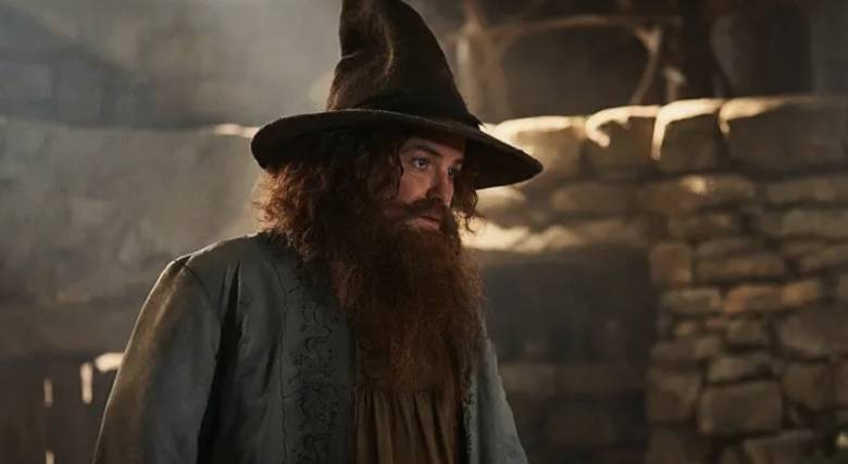 Actor Rory Kinnear dressed in wizards hat with long hair and a beard, he is wearing a grey overshirt and standing in the foreground with a brick wall in the background.