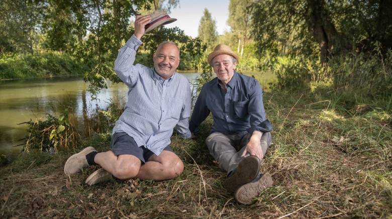 Bob Mortimer and Paul Whitehouse sitting on grass on embankment with river behind them. Bob is lifting his fishing hat and smiling while Paul sits beside him smiling