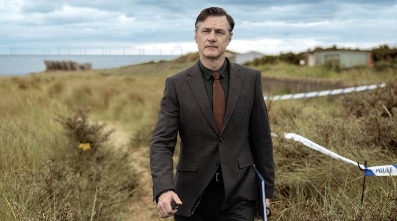 David Morrissey (Ian St Clair) wears a suit and walks along a coastal path holding a notebook and pen. Police tape cordons-off the overgrown area beside him.
