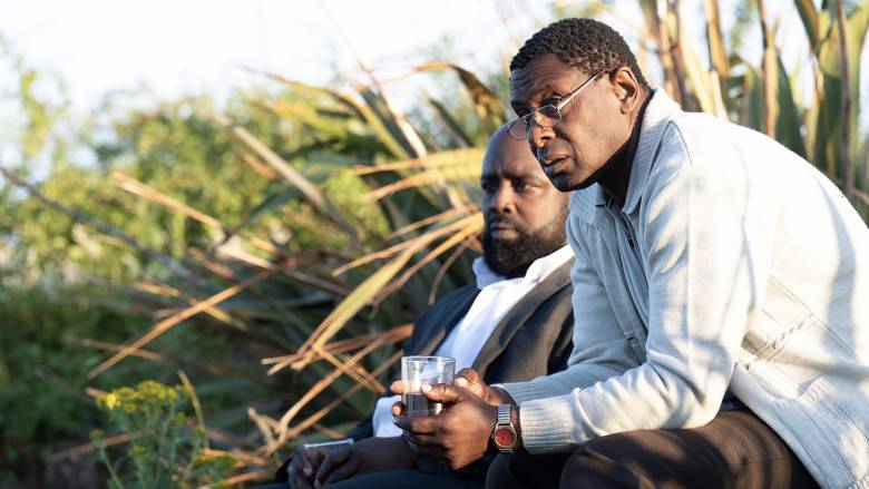 Michael Balogun (Harry Summers) and David Harewood (Denis Bottomley) sit side-by-side outdoors with green shrubs nearby. Both look at something happening off-screen. 