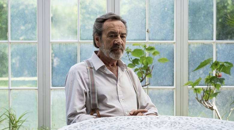 Robert Lindsay (Franklin Warner) sits at a table in a conservatory. A green garden is visible out the glass walls of the room. 
