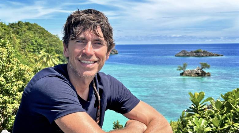 Simon Reeve smiling with blue sea and greenery behind him. the sky is blue and sunny.