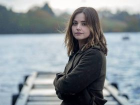 Ember (Jenna Coleman) looks to camera and stands in front of a jetty that juts out over a large body of water in the background.