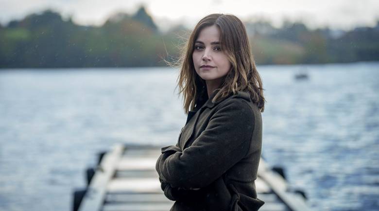 Ember (Jenna Coleman) looks to camera and stands in front of a jetty that juts out over a large body of water in the background.