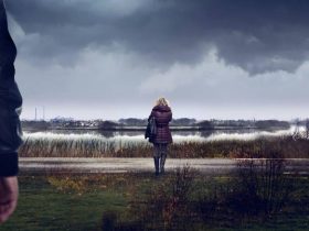 Key art for Sambre – Anatomy of a Crime. A unidentifiable person stands close to camera, facing towards the other person in the image. A woman stands a short distance away, by a roadside with her back to the camera. A bus shelter is in the corner of the scene. Grey clouds hang overhead.