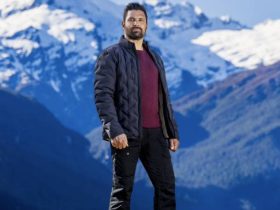 Host of The Summit Manu Bennett standing on a clifftop with mountains in the background behind him.