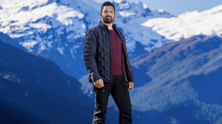 Host of The Summit Manu Bennett standing on a clifftop with mountains in the background behind him.